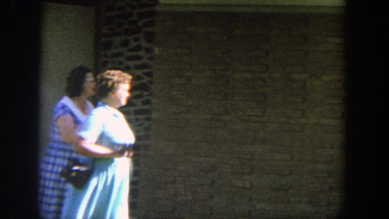 Two women in blue dresses walk side by side past a brick wall.