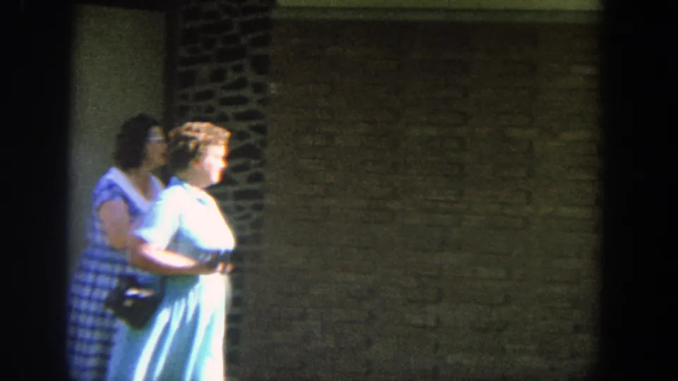 1964 Wisconsin 8mm frame with two adult women walking together beside a tan brick building with decorative dark stonework.