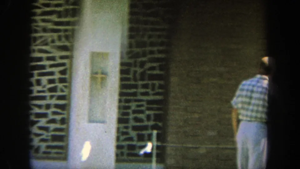 1964 Wisconsin 8mm frame with a partial adult figure at the right edge near a tan brick wall, white doorway, and dark stone trim.