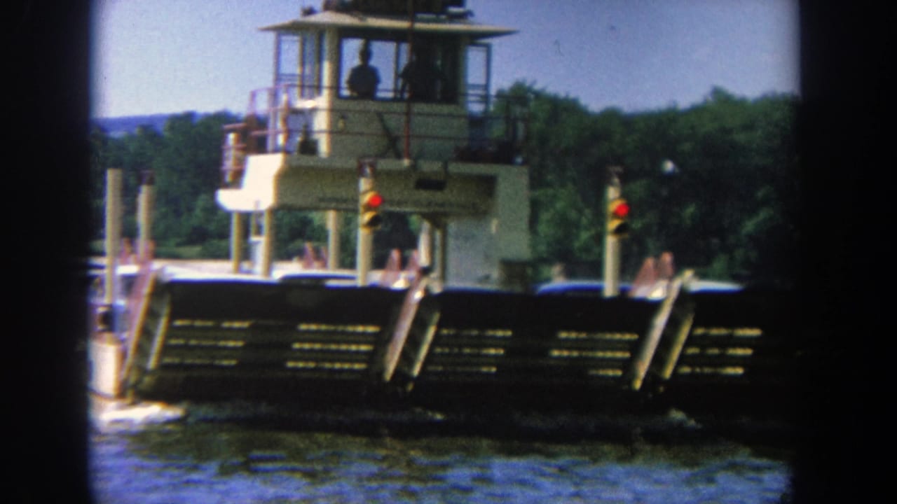 Ferry moving on a river with a raised control cabin and wooded shoreline behind it.