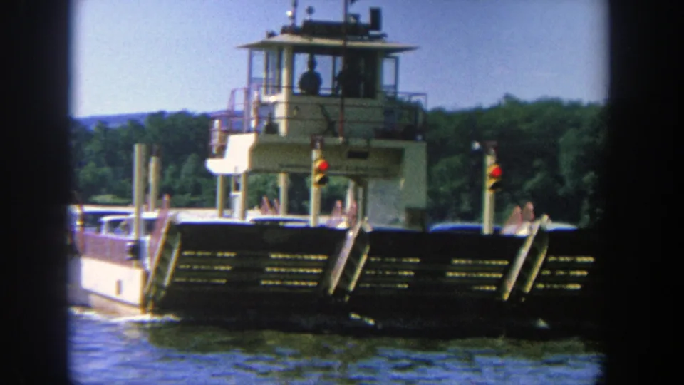 1964 Wisconsin home-movie frame of a ferry crossing calm water, with the pilot house and deck details visible against a wooded bank.