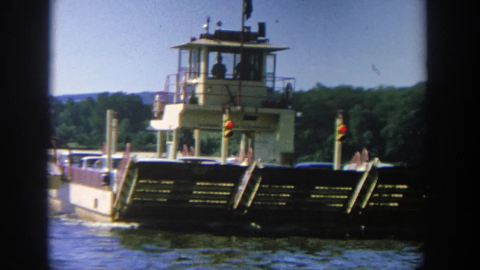 1964 Wisconsin 8mm still showing a ferry passing by, its light hull and dark bow ramp visible above rippling river water.