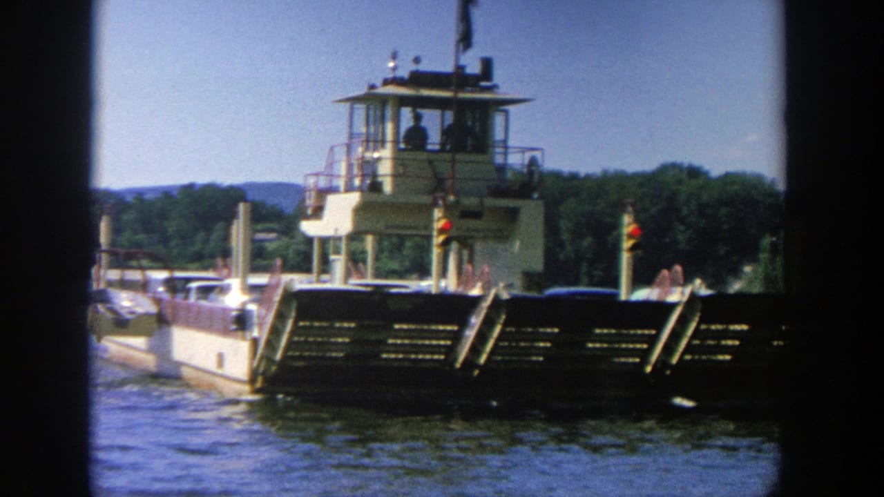 Ferry boat on calm water with pilothouse, cars, dock posts, and wooded shore.