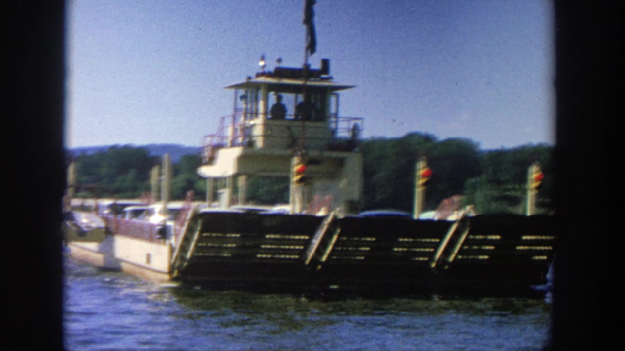 1964 Wisconsin ferry scene with a white pilothouse, open deck, cars, small passengers, and trees beyond the water.