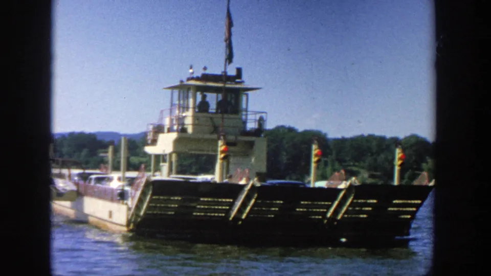 Home-movie frame from 1964 Wisconsin showing a ferry crossing calm water, with cars on deck and people visible near the railings.