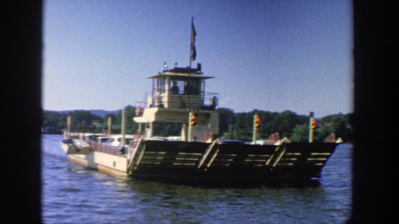Ferry boat approaching on open water with cars aboard and a wooded shoreline behind it.