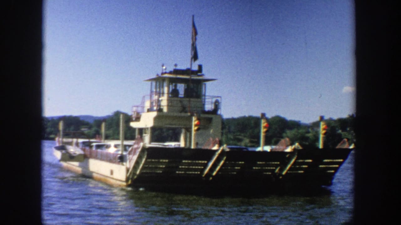 Front view of a ferry boat on water with vehicles aboard and a shoreline in the background.