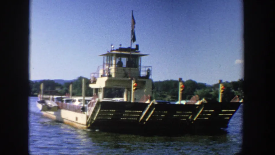 1964 Wisconsin home-movie frame showing a vehicle ferry from the front, with deck rails, cars, pilothouse, water, and distant trees.