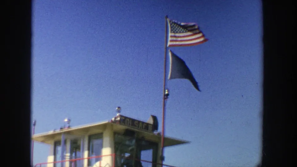 1964 Wisconsin 8mm frame focused on the ferry pilothouse, mast, American flag, blue flag, railing, and clear blue sky.