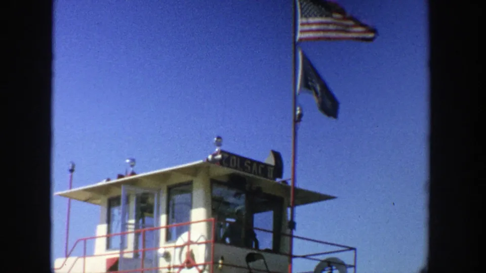 1964 Wisconsin home-movie frame of the ferry cabin roof, mast, American flag, dark blue flag, deck railing, and possible vessel lettering.
