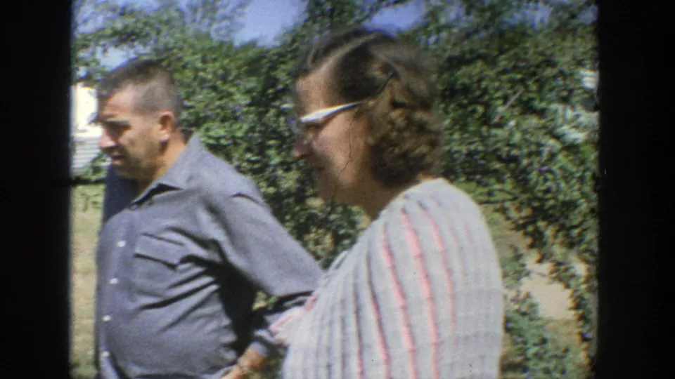 A man in a blue shirt and a woman in glasses and a striped top stand together outdoors in 1964 Wisconsin footage.
