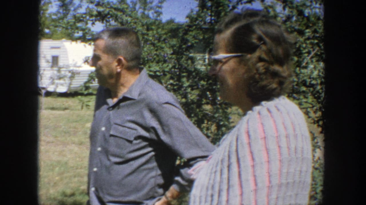 Man and woman walking past shrubs with a trailer-like structure in back.