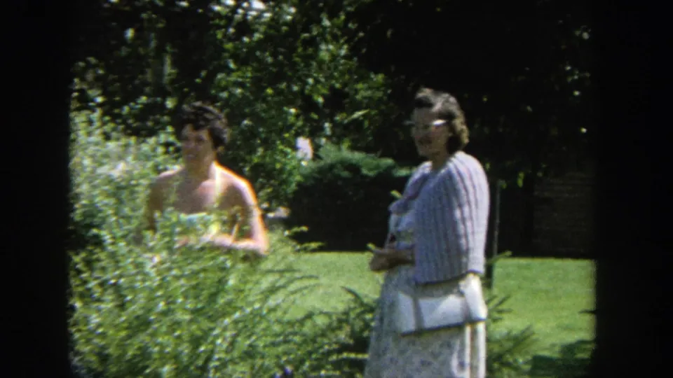 Two women stand in a green garden area in the same 1964 Wisconsin home-movie clip, one wearing a light dress and the other a striped top.