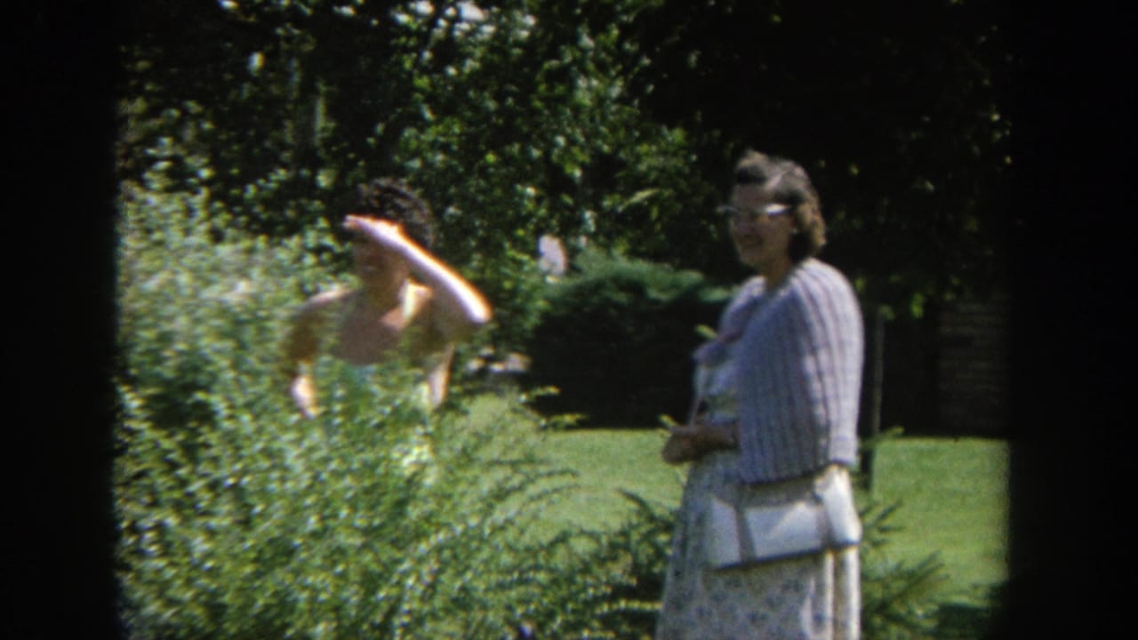 Two women in a sunny garden, one partly hidden by shrubs and one standing nearby.