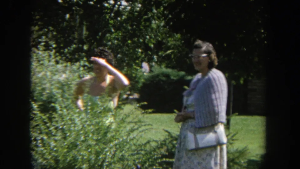 1964 Wisconsin 8mm frame showing a woman peeking through garden shrubs while another woman stands on the lawn nearby.