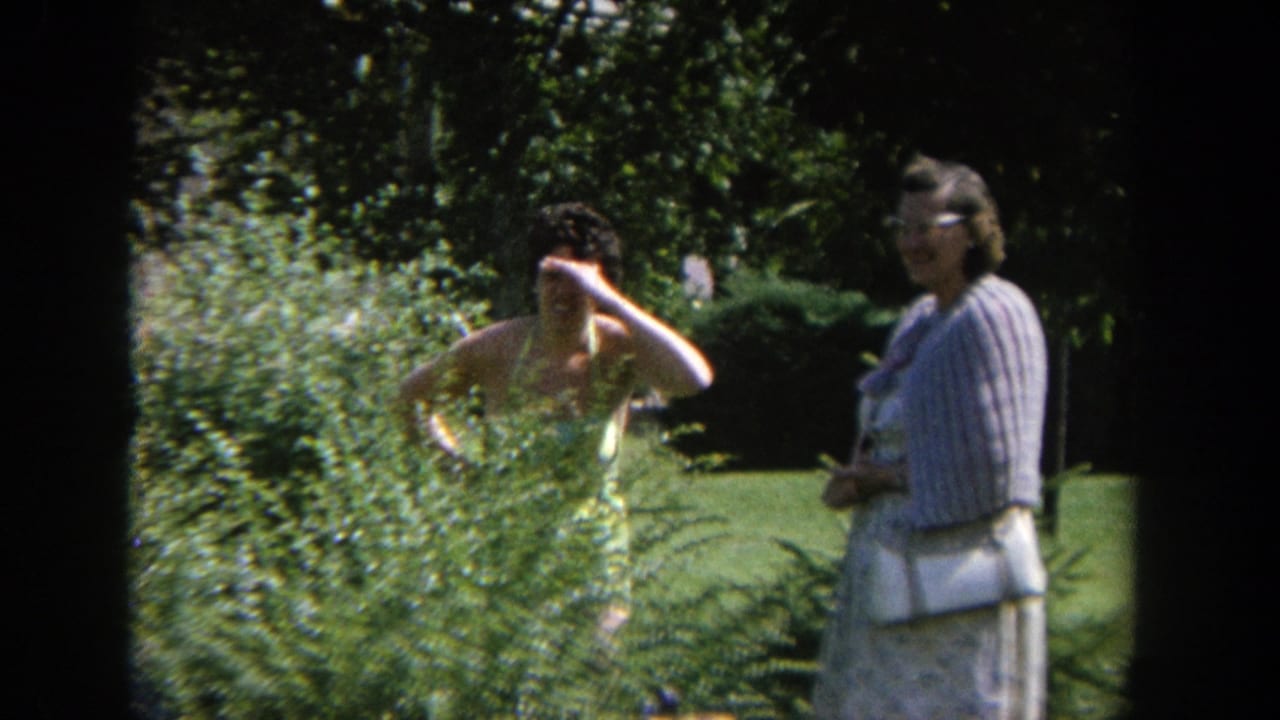 Woman in a green swimsuit shields her eyes behind shrubs as another woman stands nearby.