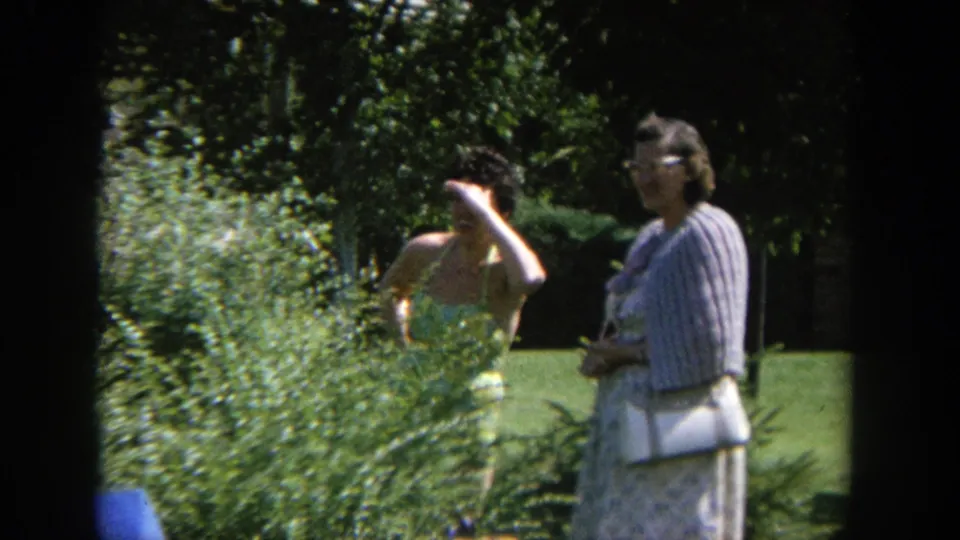 1964 Wisconsin 8mm frame of two women in a garden, one obscured by leafy shrubs and one standing in a striped cardigan.
