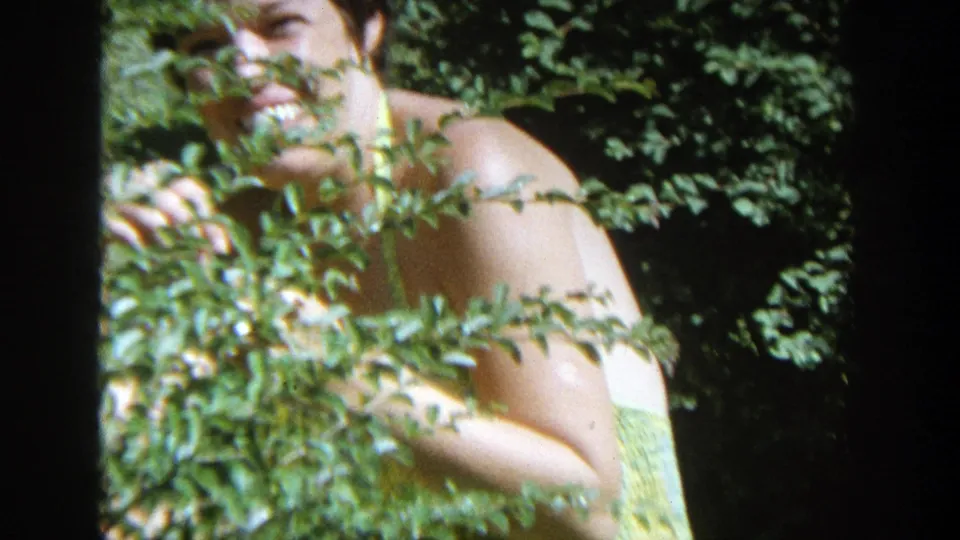 1964 Wisconsin 8mm close frame of a woman smiling from behind leafy garden branches in bright outdoor light.
