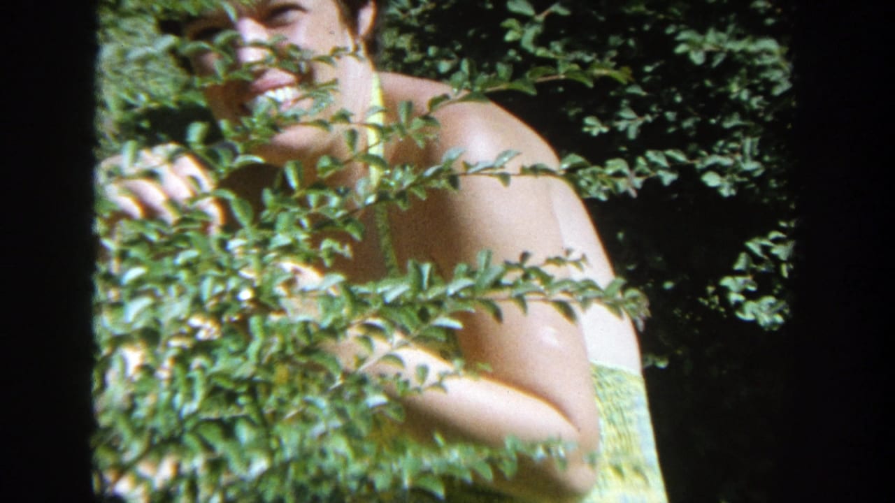 In 1964 Wisconsin home-movie footage, a woman in a yellow patterned dress smiles while partly hidden by a leafy bush.