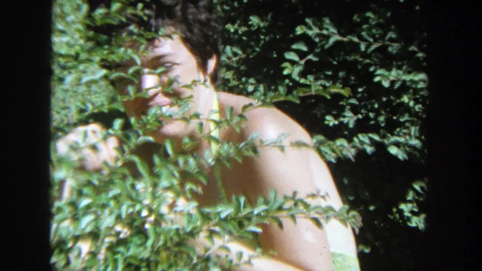 A smiling woman leans into view behind a leafy bush in a sunlit Wisconsin garden, filmed on 8mm in 1964.