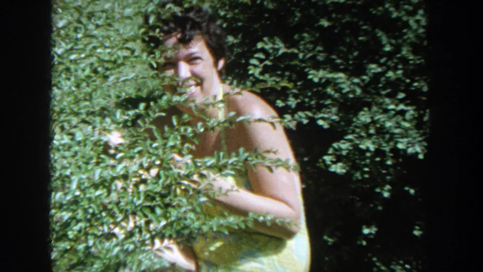 The woman smiles broadly behind dense green foliage, with her yellow dress visible in 1964 Wisconsin home-movie footage.