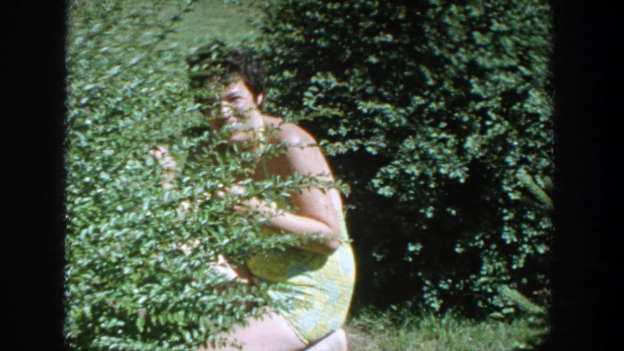 Woman in a yellow dress crouching behind a leafy hedge in a sunny garden.