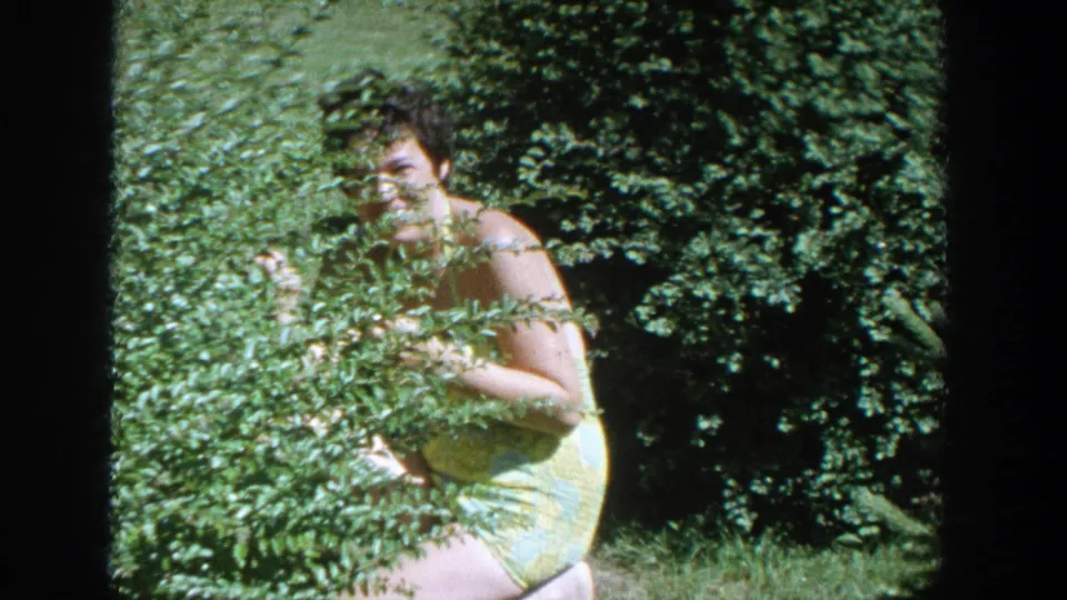 In 1964 Wisconsin home-movie footage, a smiling woman in a yellow dress peeks through dense garden foliage.
