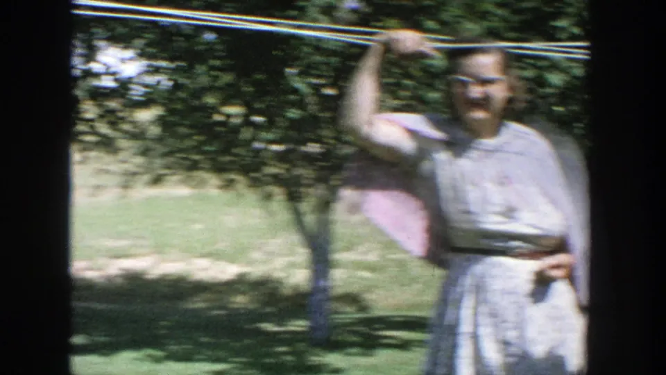 In this 1964 Wisconsin home-movie frame, a woman in a light dress and pink shawl stands by a clothesline in the backyard.