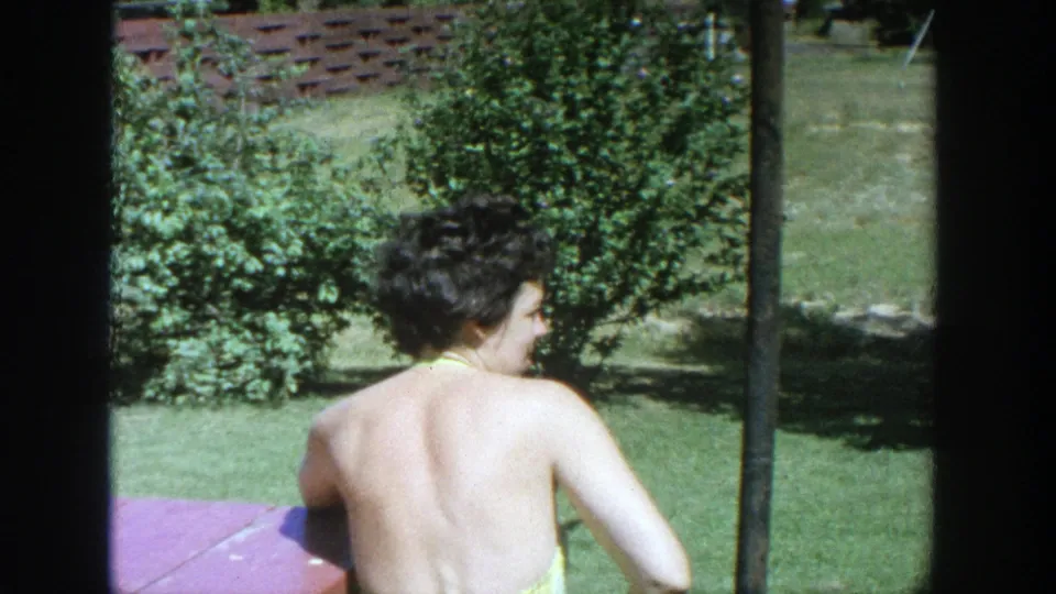 A woman seen from behind sits or turns on the lawn in a 1964 Wisconsin backyard, with shrubs and a clothesline pole nearby.