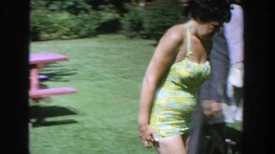 A woman in a yellow-green patterned swimsuit walks through a Wisconsin backyard past a red picnic table in 1964 footage.