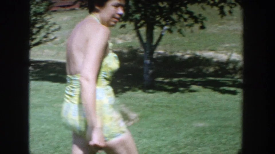 The woman in the yellow-green patterned swimsuit turns while walking across the sunny Wisconsin backyard in 1964 home-movie footage.