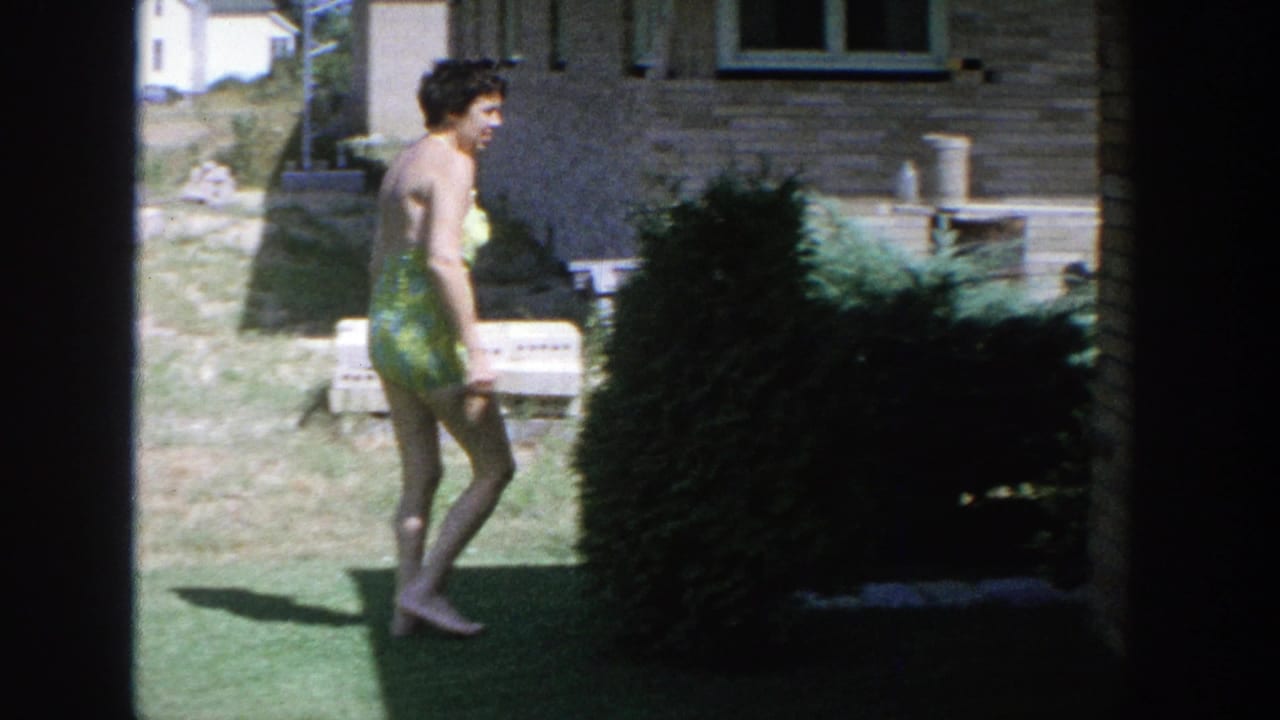 Woman in a green swimsuit walks across a sunny backyard near a brick house and shrub.