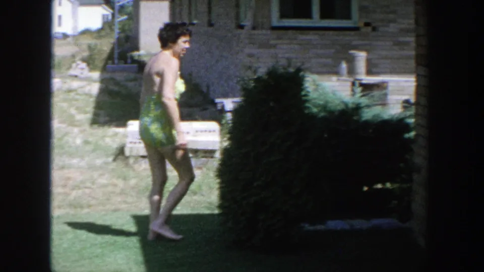 1964 Wisconsin 8mm frame showing a woman in a green patterned swimsuit walking on grass beside a brick house and large shrub.