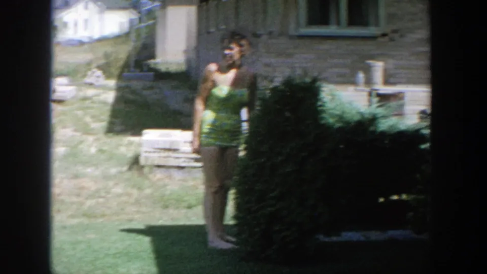 1964 Wisconsin home-movie frame with a woman smiling toward the camera in a suburban backyard beside a shrub and brick house.