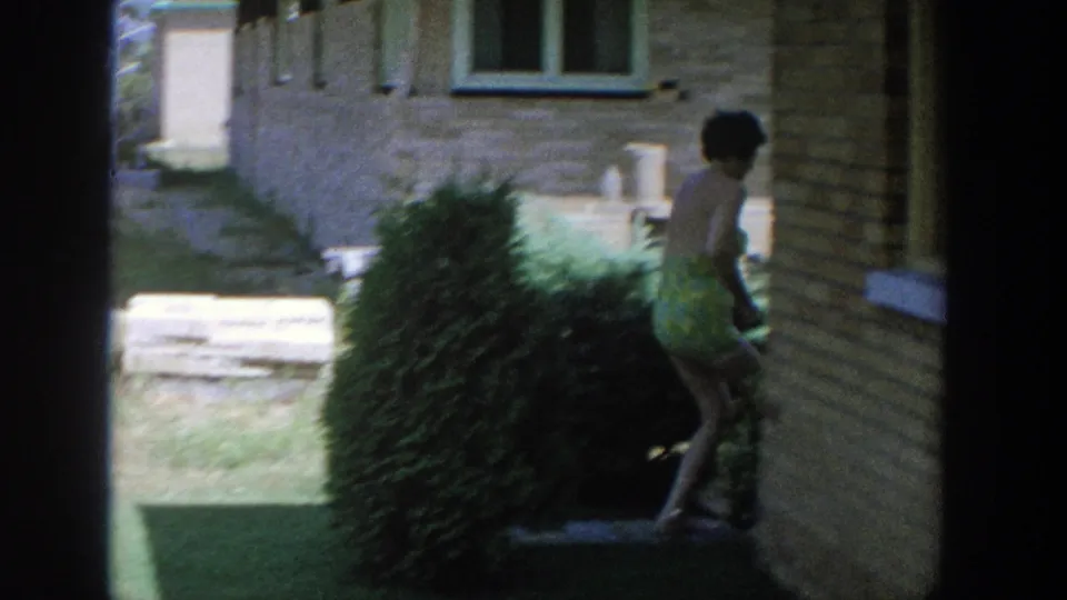 1964 Wisconsin 8mm frame showing the woman ducking behind a shrub along the side of a brick house in the backyard.