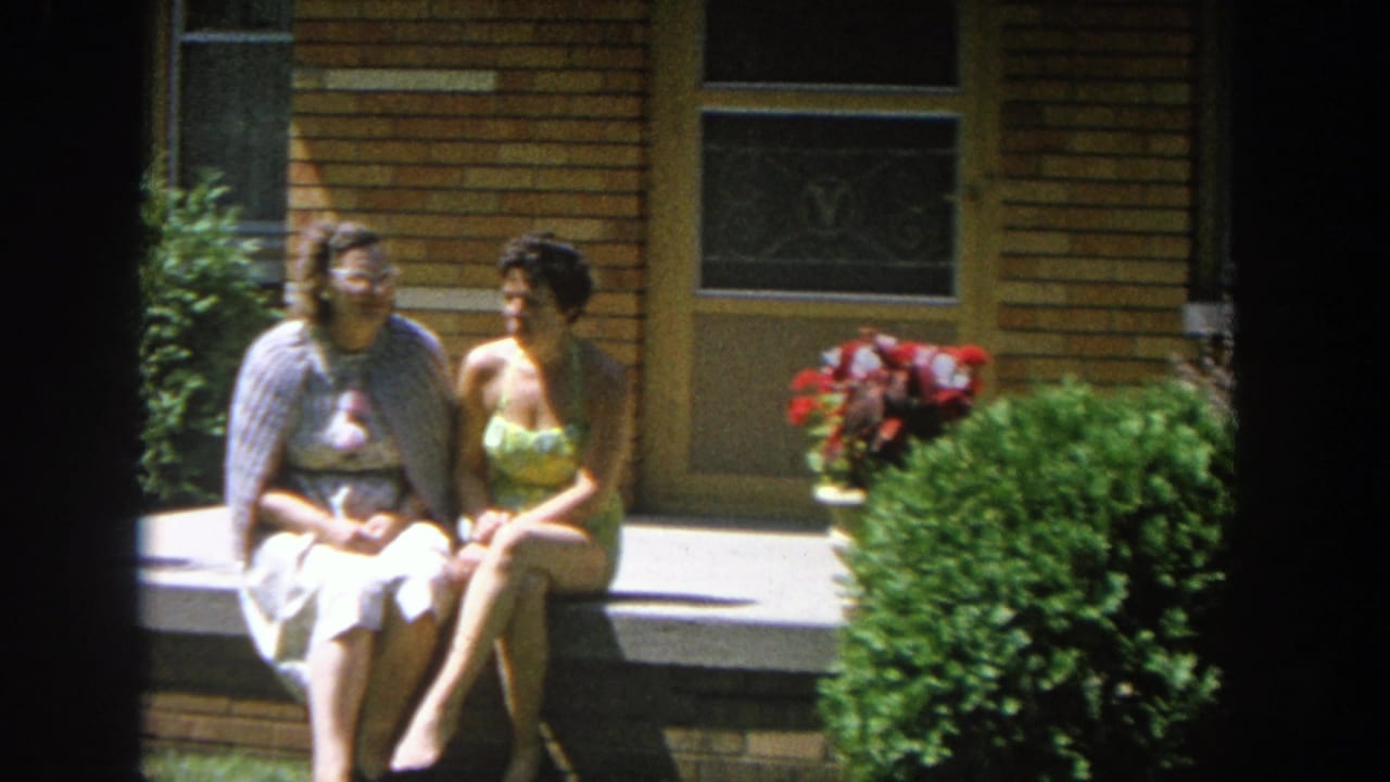 Two women sit on a concrete porch step outside a tan brick house.