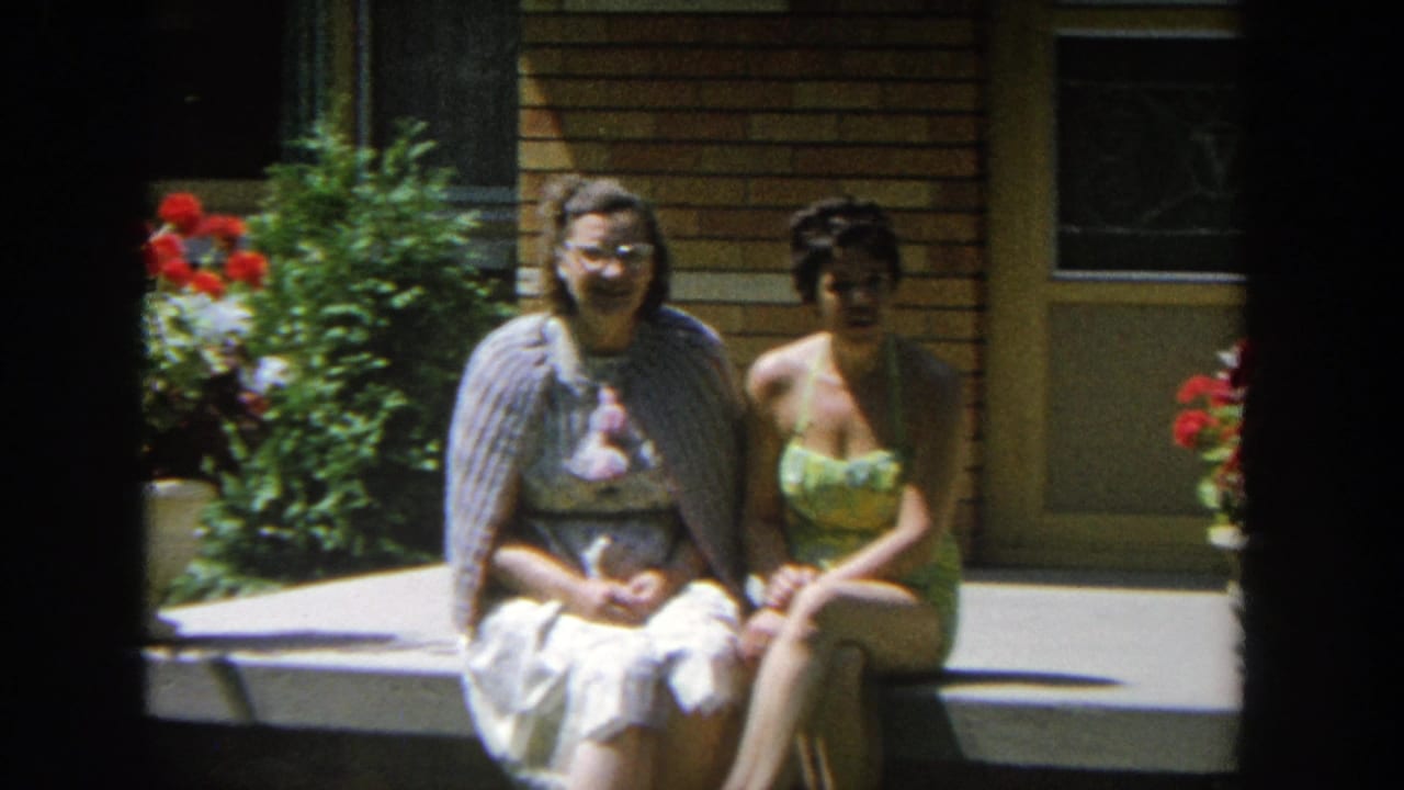 Two women face the camera while seated on a porch step by red flowers.