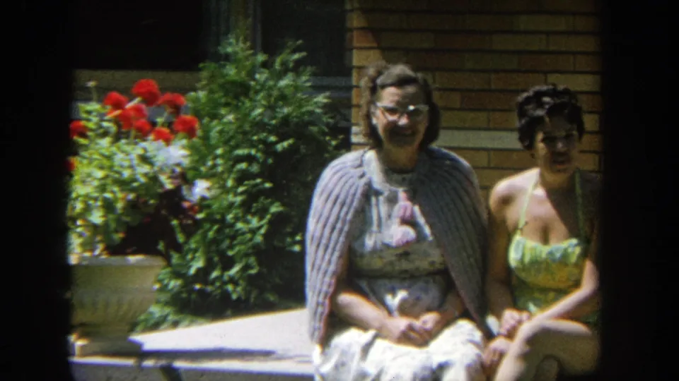 A closer 8mm frame shows the two women seated together near a planter of red flowers and green shrubs outside the tan brick house.