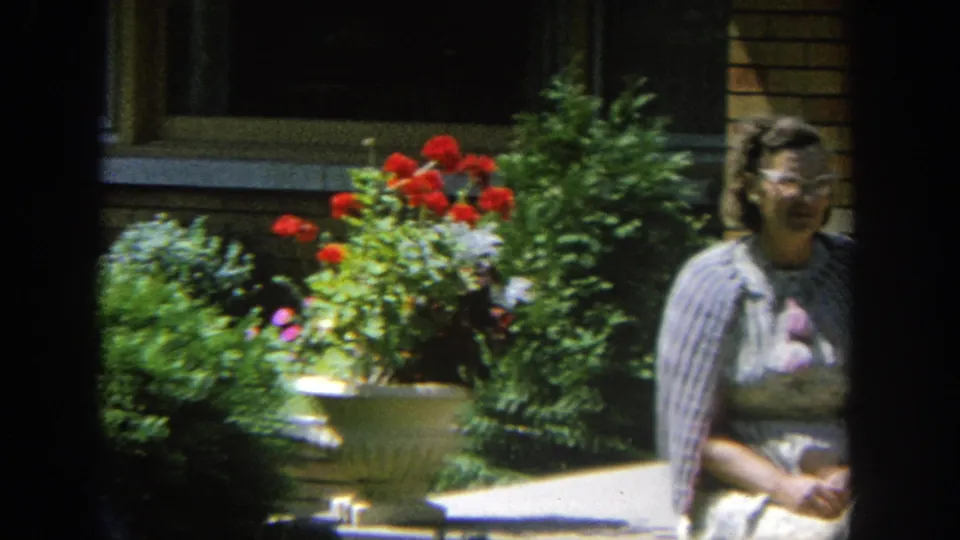 The camera drifts across the porch plants, leaving one seated woman partly visible at frame edge against the tan brick house.