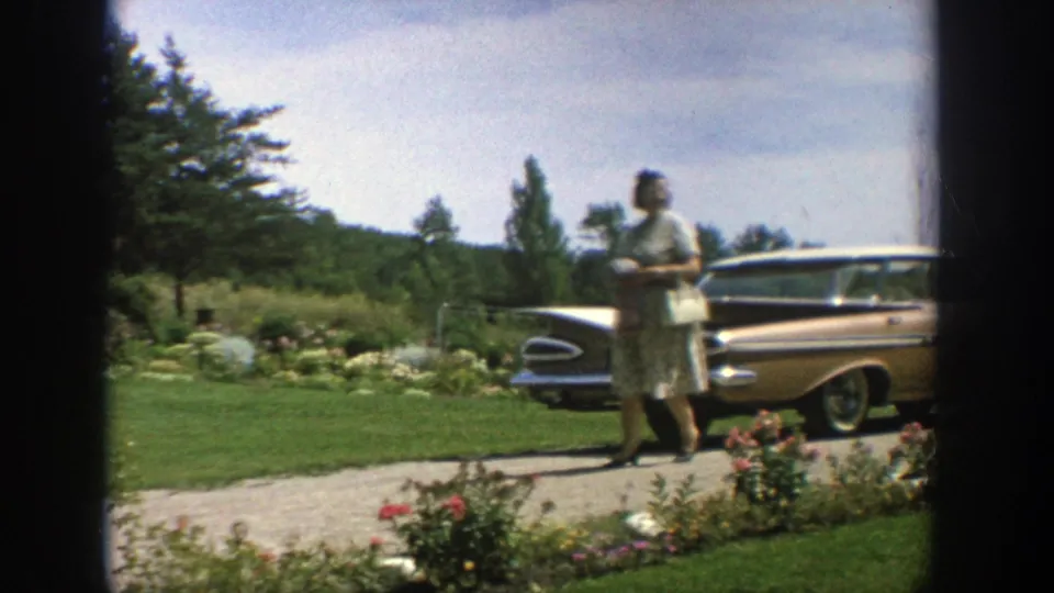 Woman in a light dress walks along a Wisconsin driveway beside a parked mid-century car, lawn, garden beds, and trees.