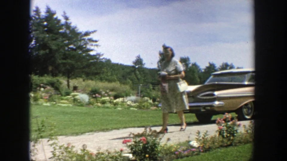 Woman in heels and a light dress stands or walks by the driveway near a parked car in a 1964 Wisconsin residential garden scene.