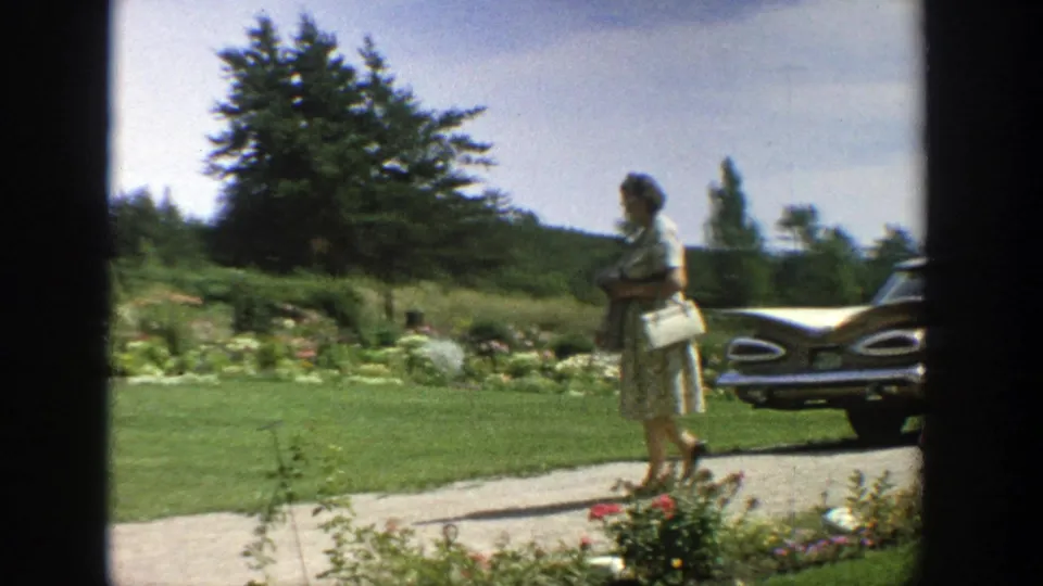 In 1964 Wisconsin home-movie footage, a woman in a patterned dress walks through a garden yard near a tailfin-era car.