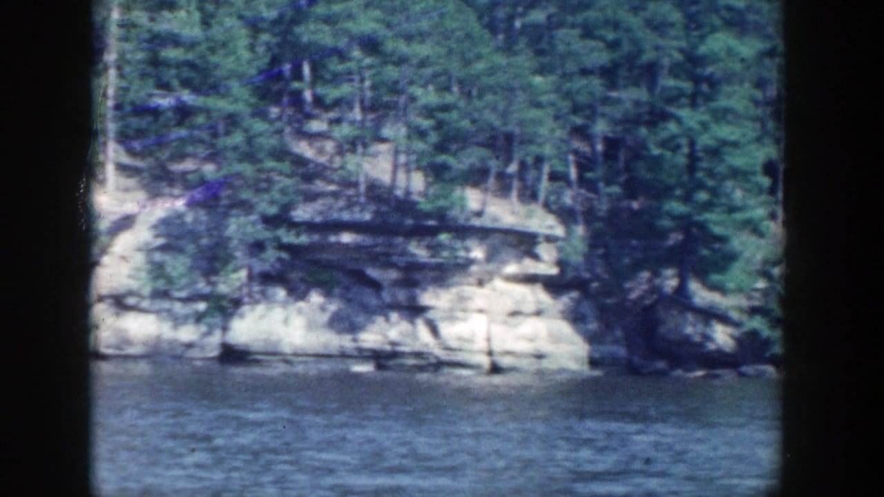 Wisconsin river shoreline with layered rock cliffs and dense trees in 1964 8mm home-movie footage.