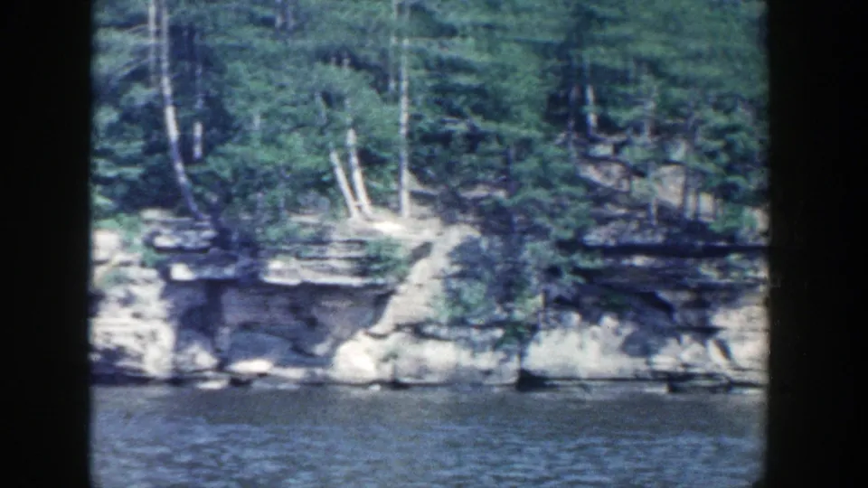 Layered rock ledges and trees line a Wisconsin riverbank in a 1964 home-movie shoreline view.