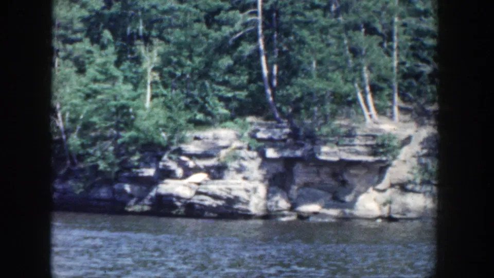 Rippling water faces a rocky, tree-lined Wisconsin shoreline in 1964 color 8mm footage.