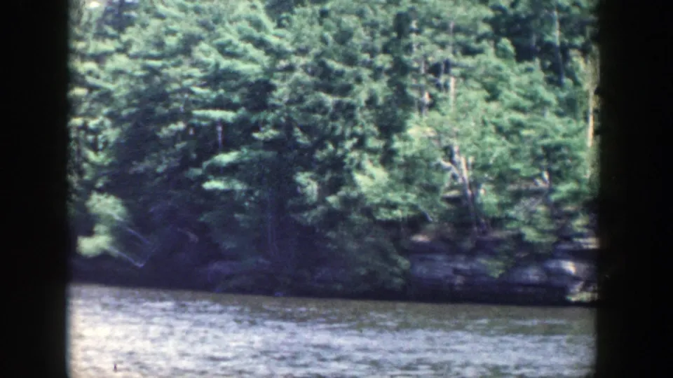 1964 Wisconsin home-movie view of a shaded wooded bank and rock edge seen from across the river.
