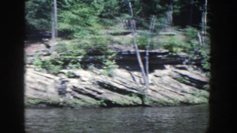 1964 Wisconsin frame of layered rock formations and small trees along a riverbank in daylight.