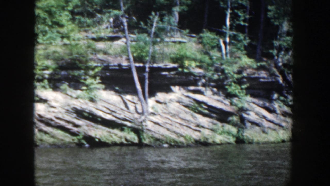 Wooded rocky shoreline across dark water, framed by black film borders.