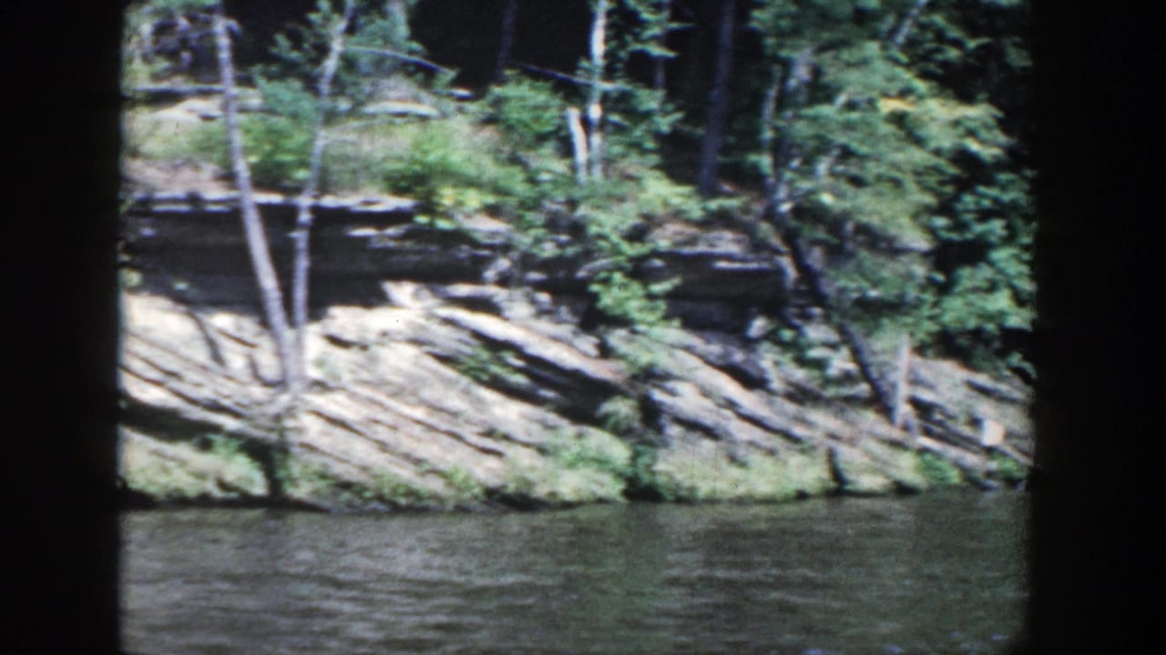 1964 Wisconsin 8mm view of a wooded rock shoreline beside dark rippling water.