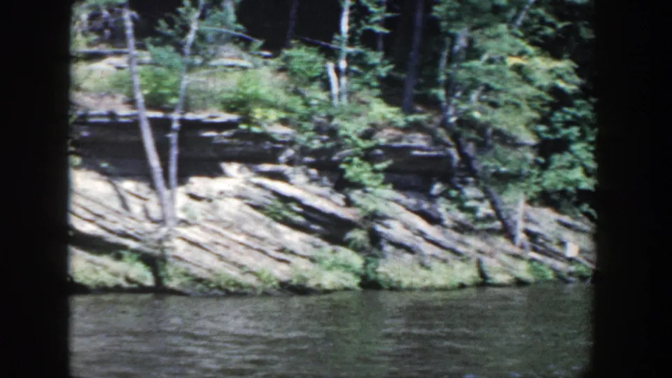 1964 Wisconsin 8mm view of a wooded rock shoreline beside dark rippling water.